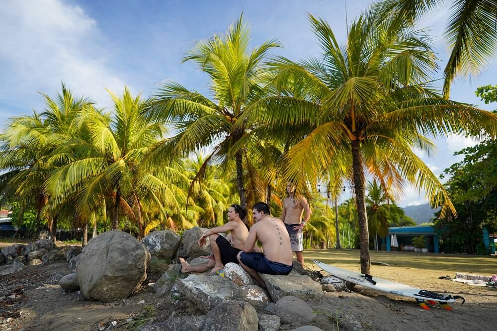 Tři mladí muži odpočívají na kamenech pod palmami na tropické pláži v Kostarice